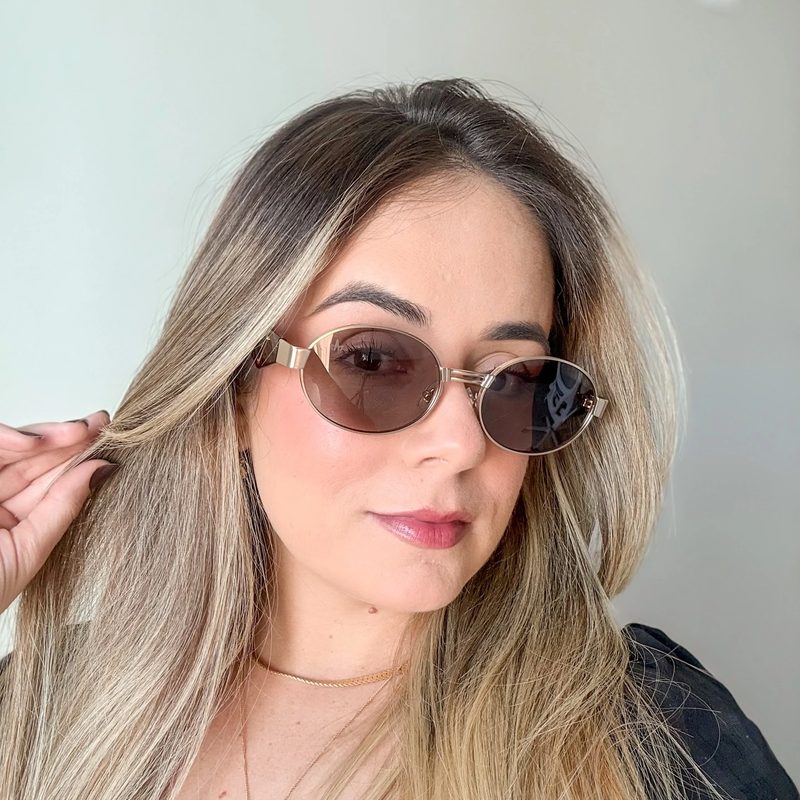 Cliente loira usando óculos de sol ovais dourados da Magic Sunglass, colar dourado, fundo neutro