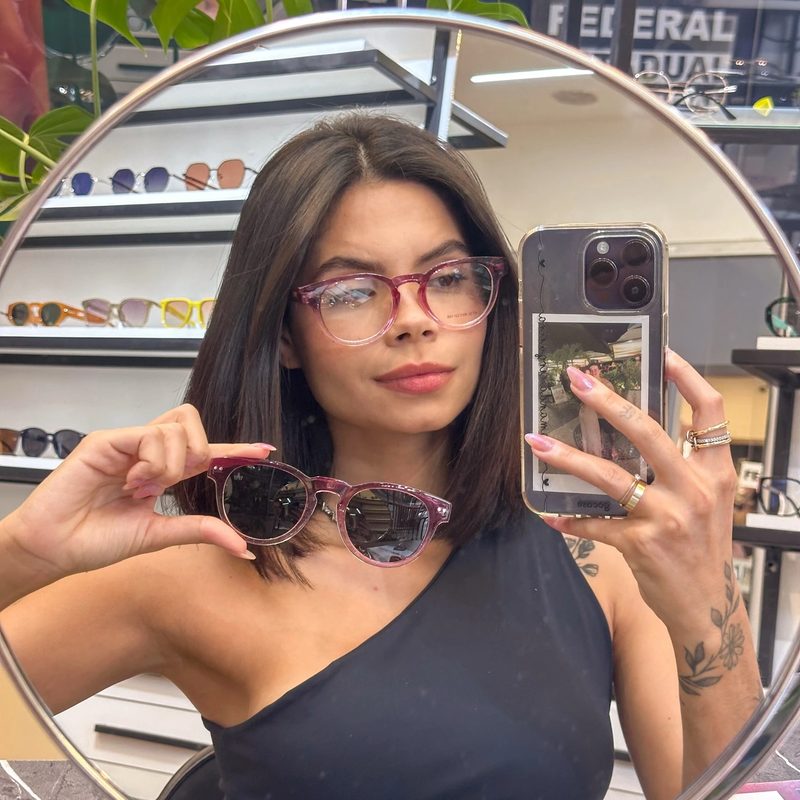 Cliente se fotografando em espelho redondo da ótica, comparando armação de grau rosa e óculos de sol rosa da Magic Sunglass