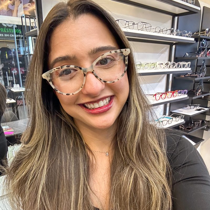 Cliente sorridente usando armação de grau mesclada em tons neutros, dentro da ótica com prateleiras de óculos ao fundo