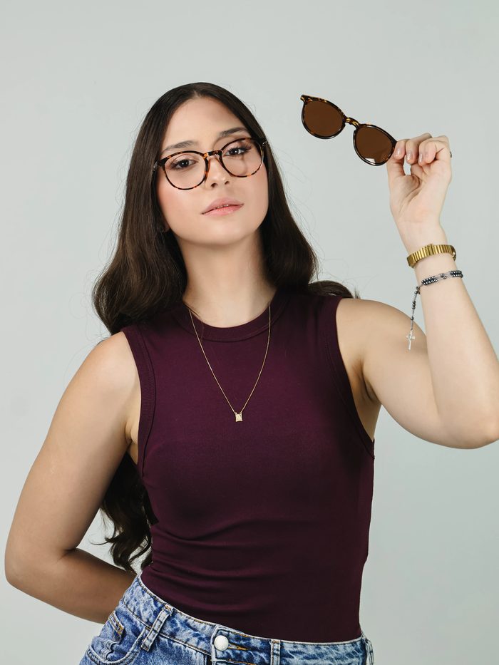 Mulher usando armação de grau, segurando óculos de sol de acetato tartaruga levantados acima da cabeça