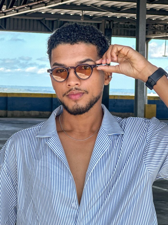 Homem usando óculos de sol redondos com lentes âmbar, ajustando a armação com a mão, em terraço com vista para o mar