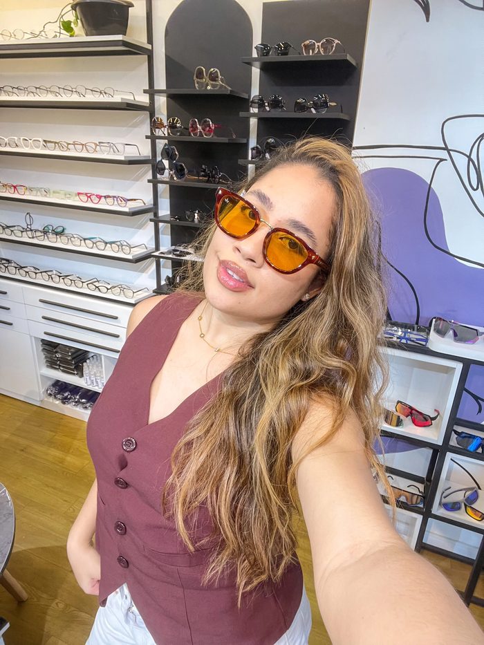 Mulher fazendo selfie usando óculos de sol com lentes laranja em loja de óticas, com prateleiras de armações ao fundo