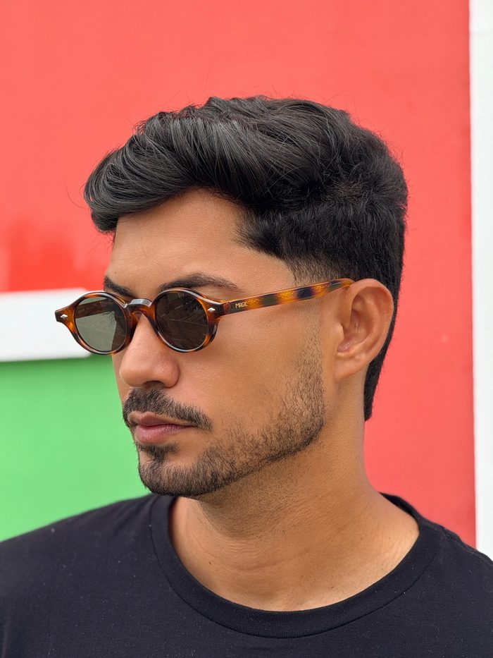 Homem de perfil usando óculos de sol redondos de armação tartaruga com a marca Magic, fundo vermelho e verde